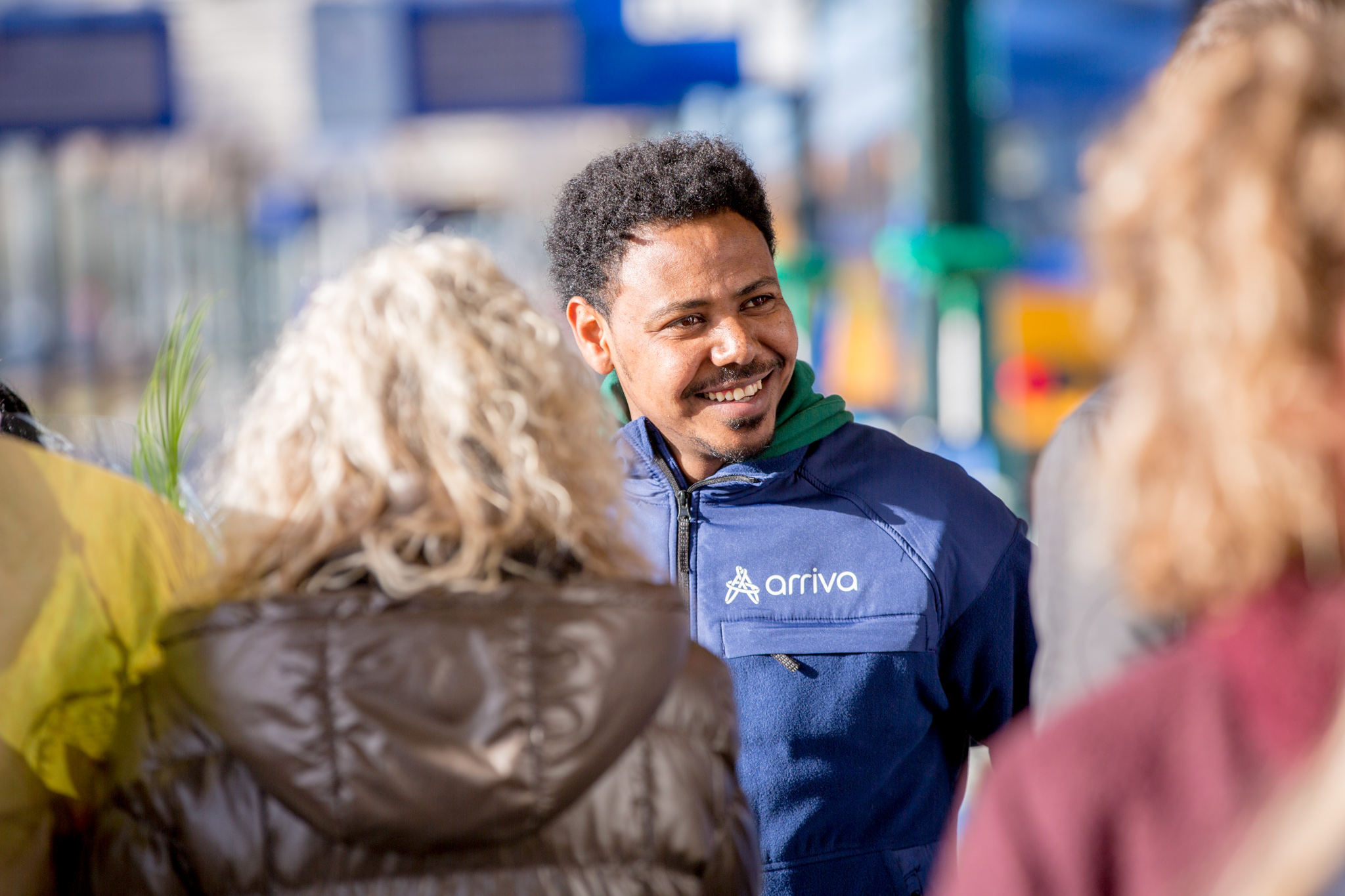 Een vrolijke man in een blauw Arriva uniform lacht naar mensen, de zon schijnt.