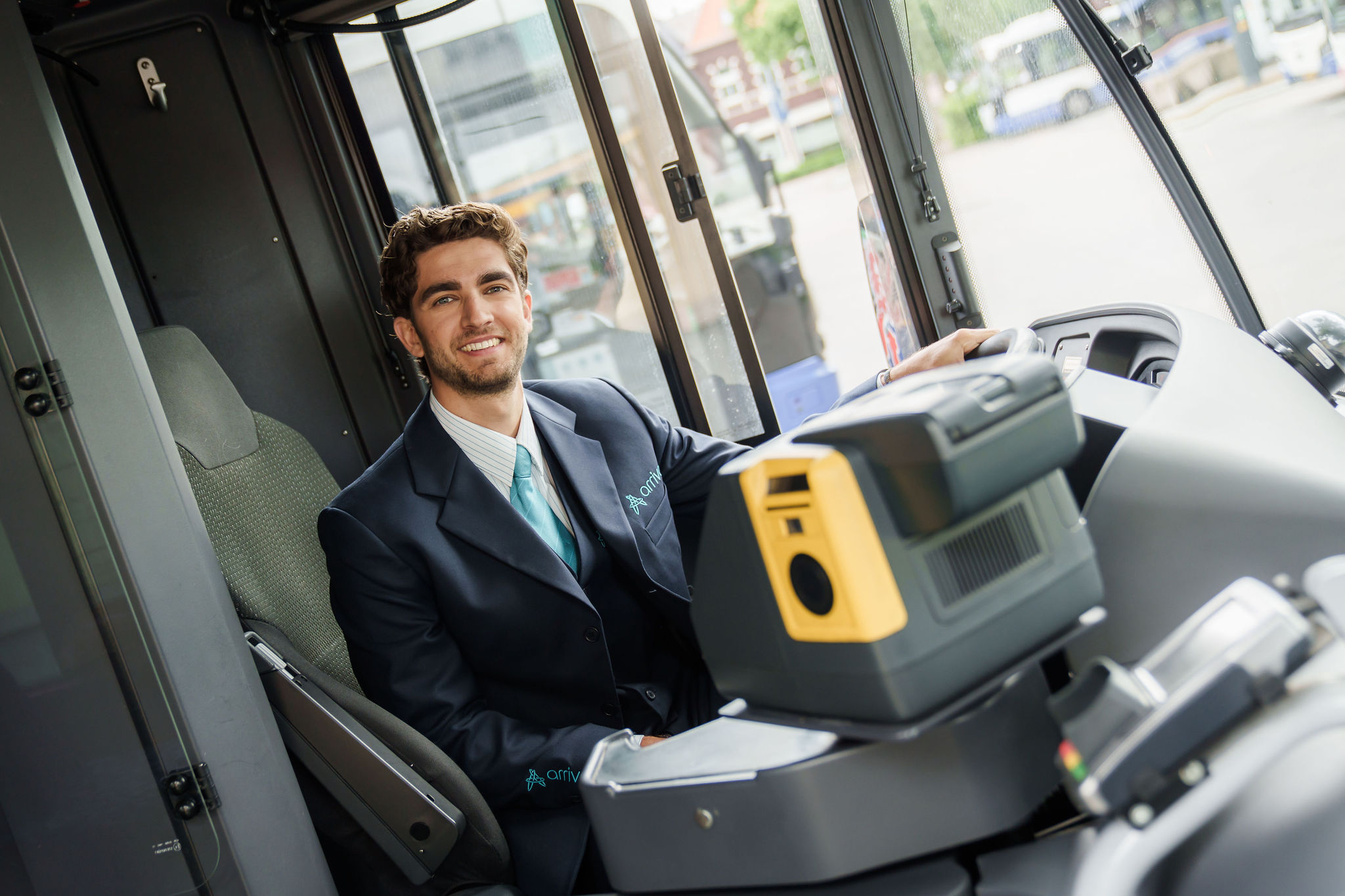 Een lachende buschauffeur in Arriva-uniform zit achter het stuur van een bus en kijkt naar de camera.