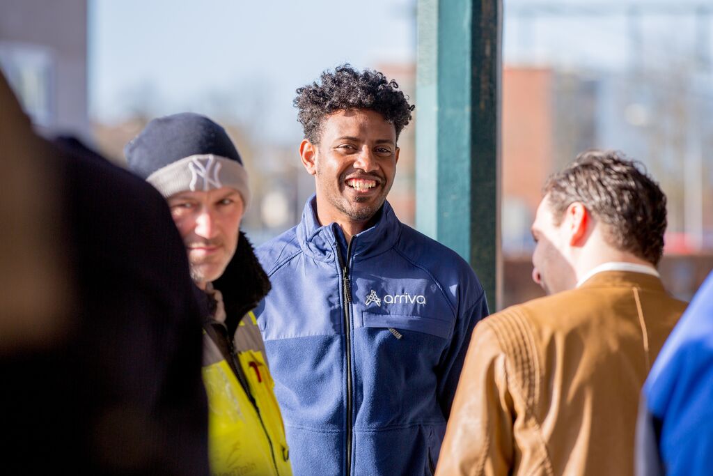 Een servicemedewerker van Arriva met zwart haar lacht naar de camera, om hem heen staan reizigers.