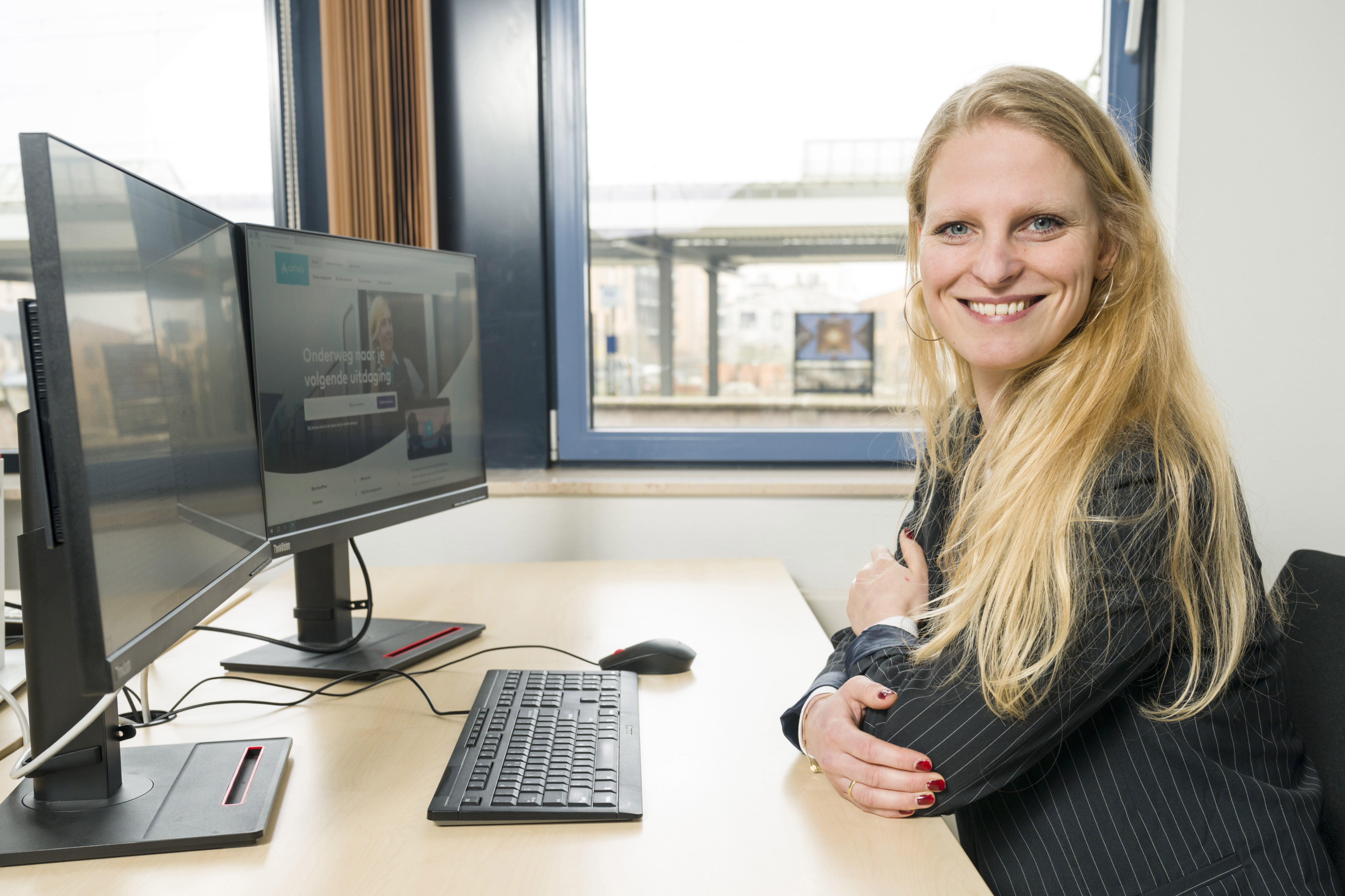 Vrouw in kantoor glimlacht naar camera, computerscherm met website Arriva zichtbaar, groot raam toont uitzicht op treinstation.