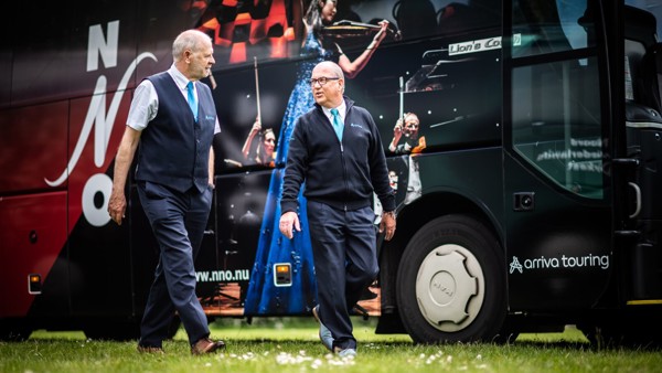 Twee mannelijke Arriva chauffeurs in uniform lopen langs een Arriva Touring bus, terwijl ze zich op een grasveld bevinden. Twee mannelijke Arriva chauffeurs in uniform lopen langs een Arriva Touring bus, terwijl ze zich op een grasveld bevinden.