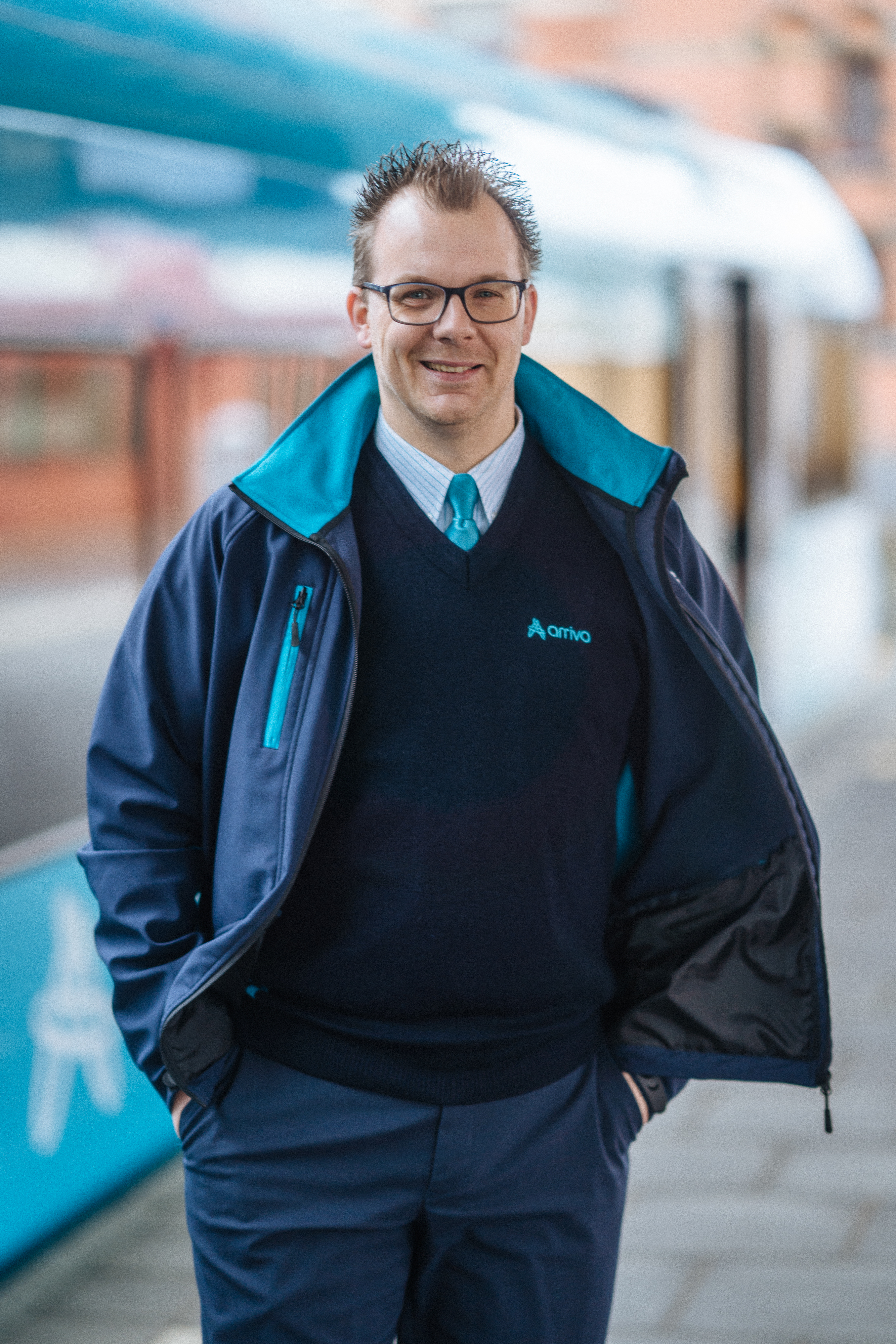 Machinist Frank staat lachend in Arriva-uniform op het perron, vaag in de achtergrond zie je een blauw-witte Arriva trein.