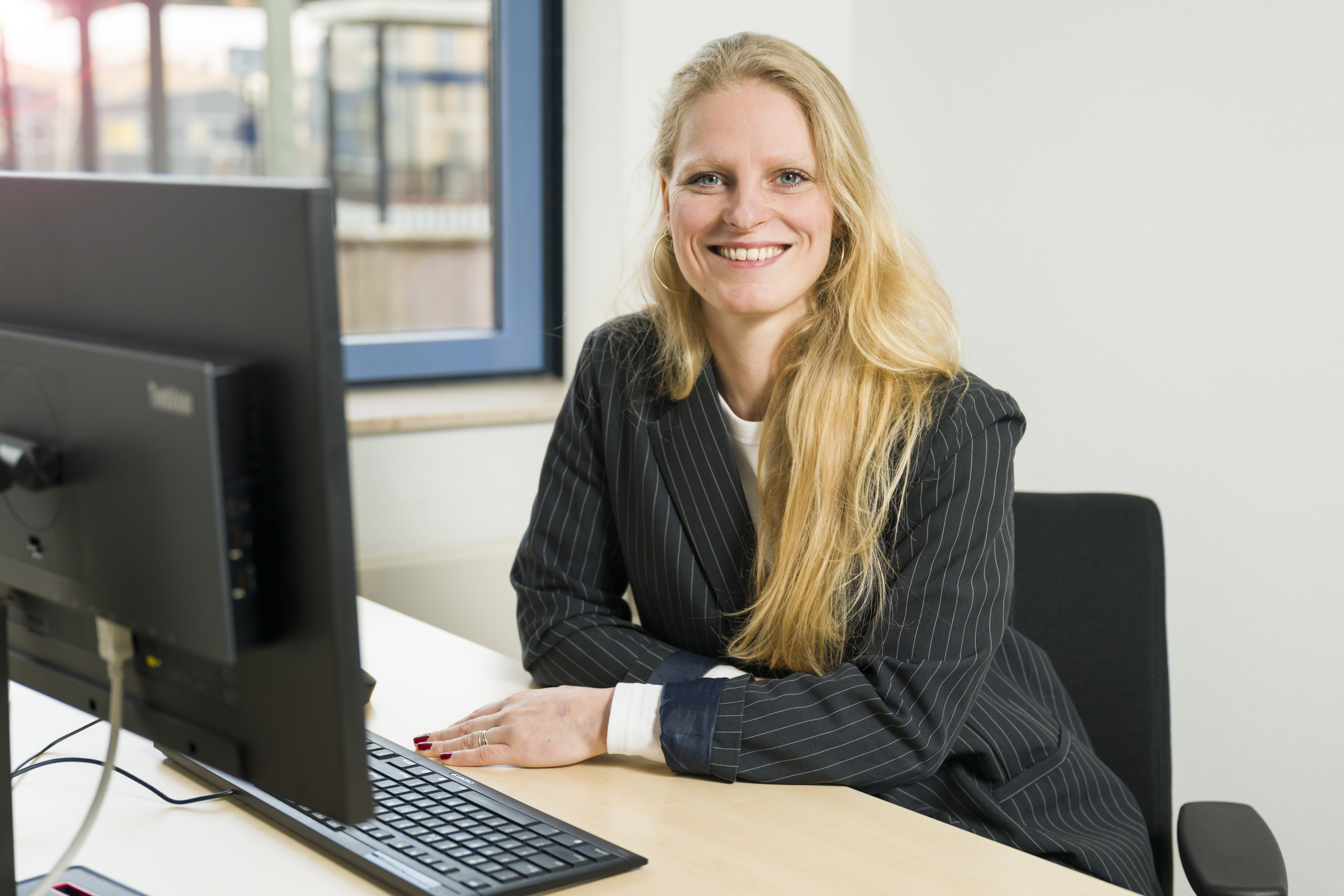 Vrouw achter computer in kantoor, glimlacht naar camera in professionele kleding. Raam biedt uitzicht op trein en stadsgebouwen bij openbaar vervoer.