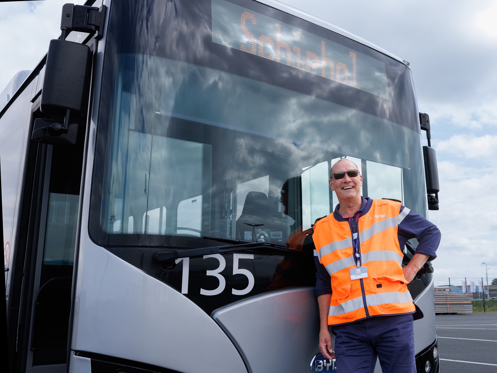 Buschauffeur in oranje hesje staat lachend voor Arriva Touringbus met Schiphol erop.