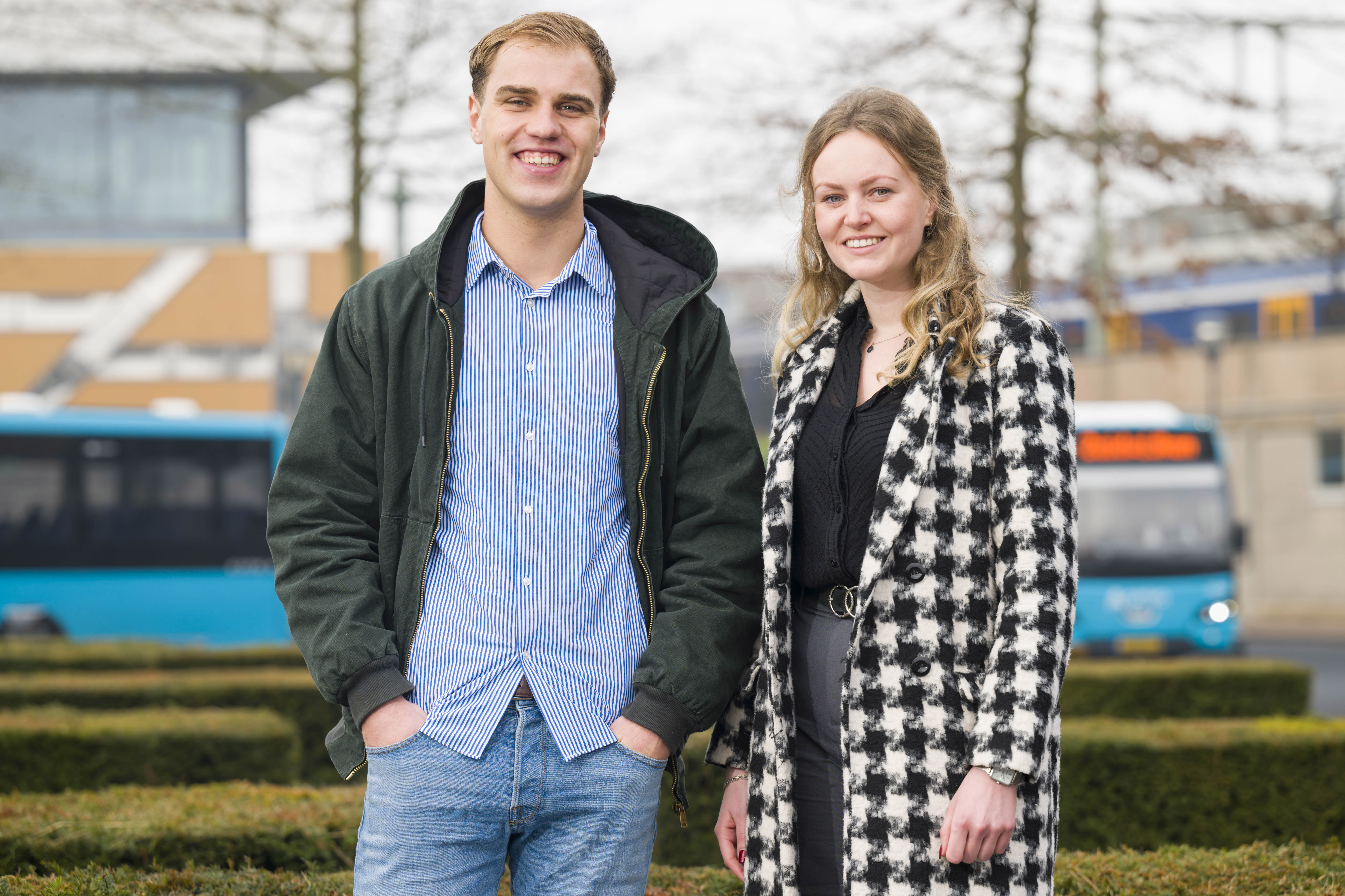 Man en vrouw buiten, glimlachend naar camera; hij in casual jas en gestreept shirt, zij in geruite jas, tegen stedelijke achtergrond met bus- en treinstation.