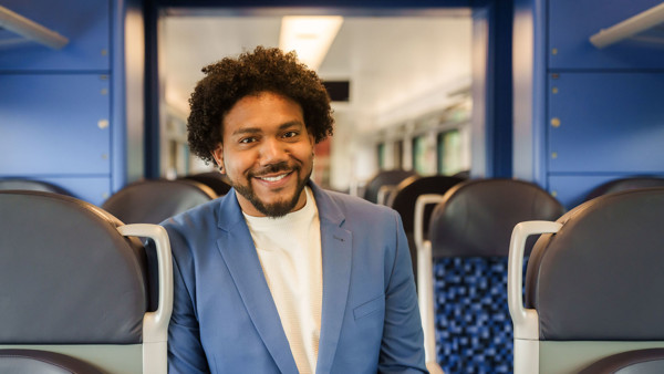 Een teammanager van Arriva staat tussen stoelen in de trein en lacht naar de camera. Een teammanager van Arriva staat tussen stoelen in de trein en lacht naar de camera.