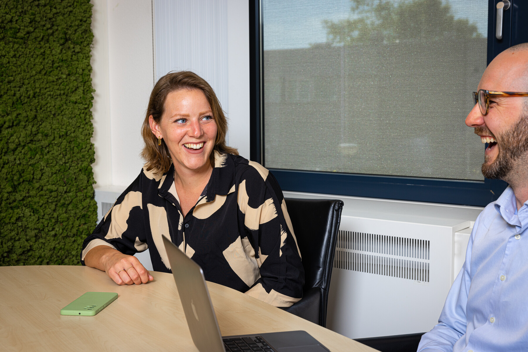 Een lachende vrouw en man zitten aan een tafel. Op tafel liggen een telefoon en een laptop.