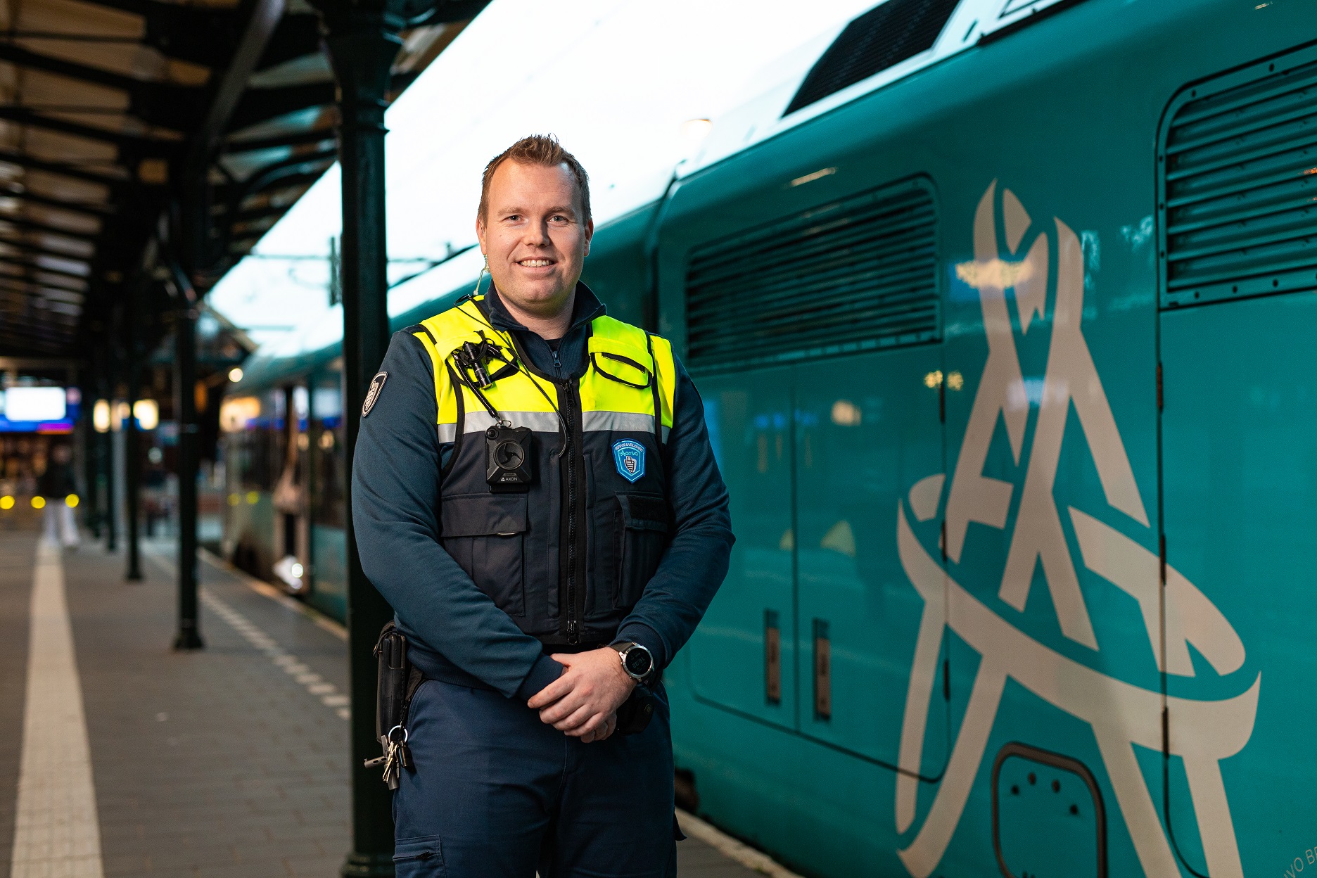 Een Steward/Boa OV staat in uniform en lachend op het perron, naast hem staat een blauwe Arriva-trein.