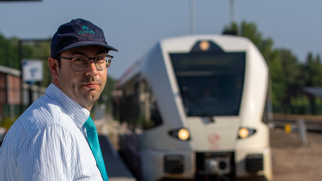 Machinist Rob kijkt recht in de camera met een pet en een bril op. Op de achtergrond staat een Arriva-trein. Machinist Rob kijkt recht in de camera met een pet en een bril op. Op de achtergrond staat een Arriva-trein.