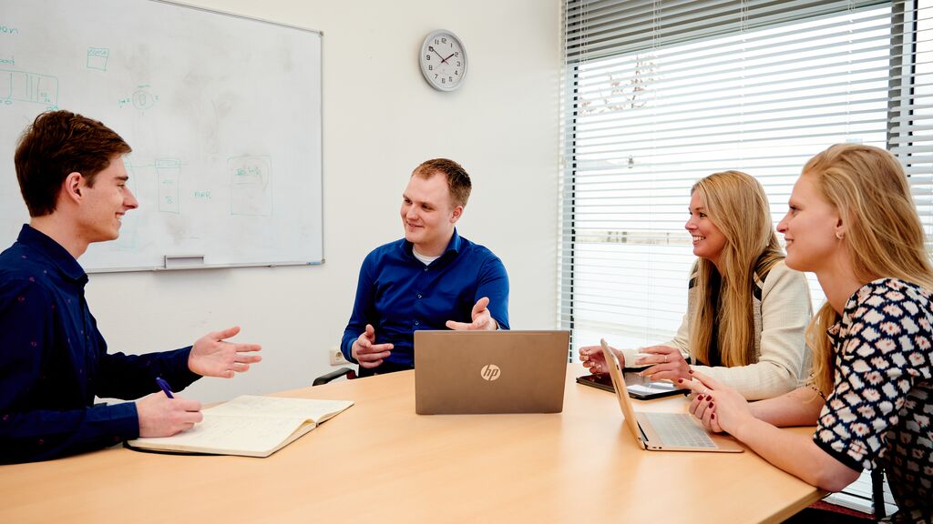 Vier jonge collega's van Arriva zitten aan een ronde tafel met laptops en vergaderen lachend met elkaar.