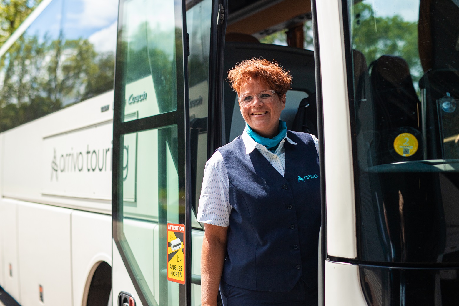 Buschauffeur Marijke staat lachend in de deuropening van haar bus waarop duidelijk 'Arriva Touring' staat.