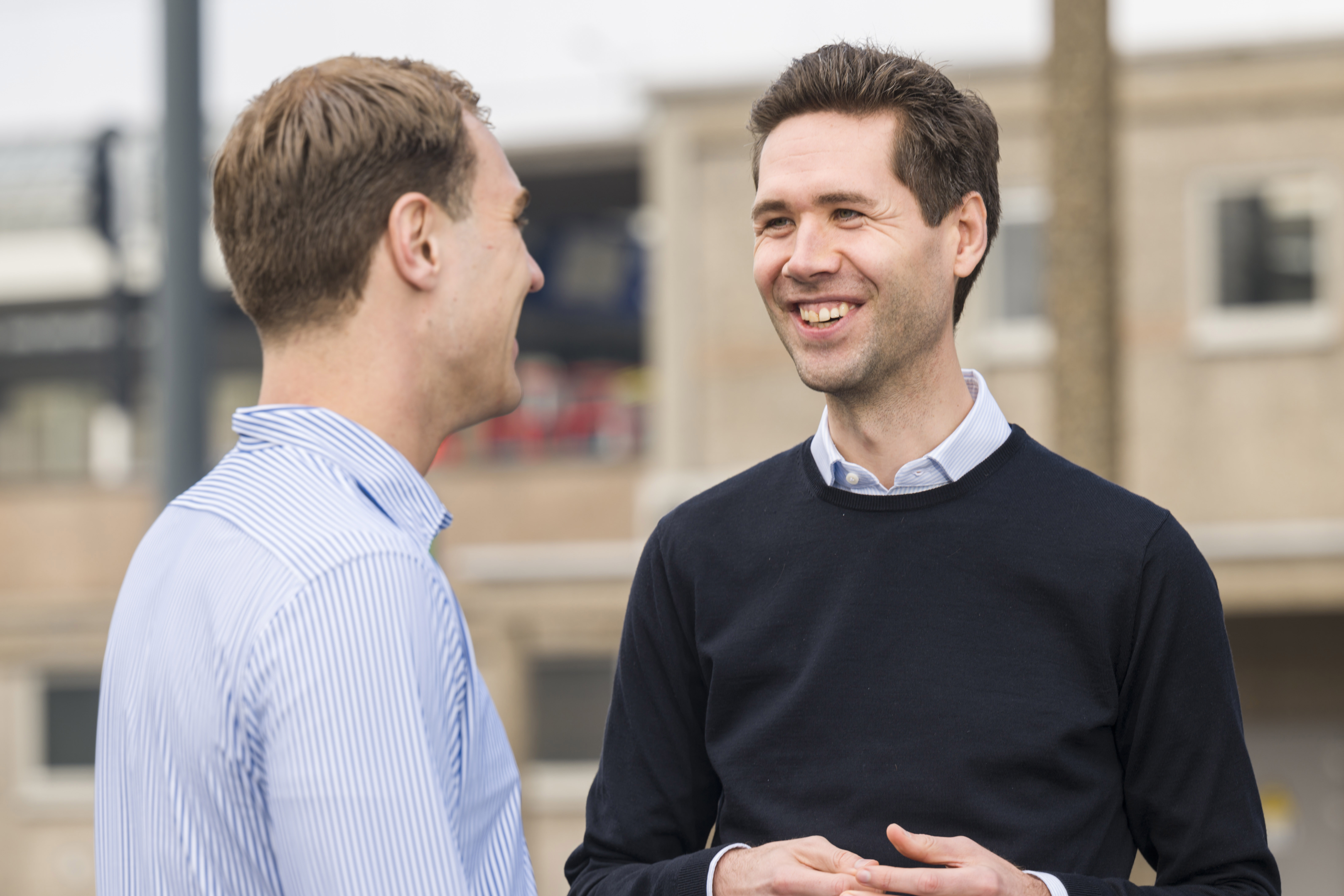 Twee mannen voeren buiten een gesprek; één gebaart actief, de ander luistert aandachtig. Op de achtergrond is een vaag stedelijk station.