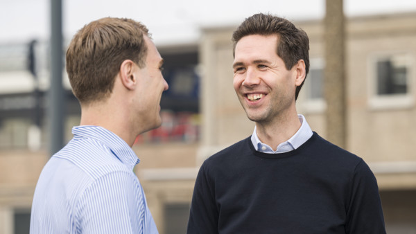 Twee mannen voeren buiten een gesprek; één gebaart actief, de ander luistert aandachtig. Op de achtergrond is een vaag stedelijk station. Twee mannen voeren buiten een gesprek; één gebaart actief, de ander luistert aandachtig. Op de achtergrond is een vaag stedelijk station.