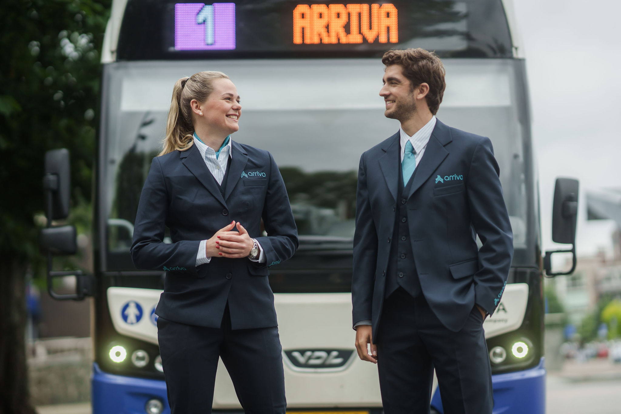 Een lachende man en vrouw in Arriva-uniforms staan voor een bus met het Arriva-logo en lijnnummer 1 zichtbaar op de achtergrond.