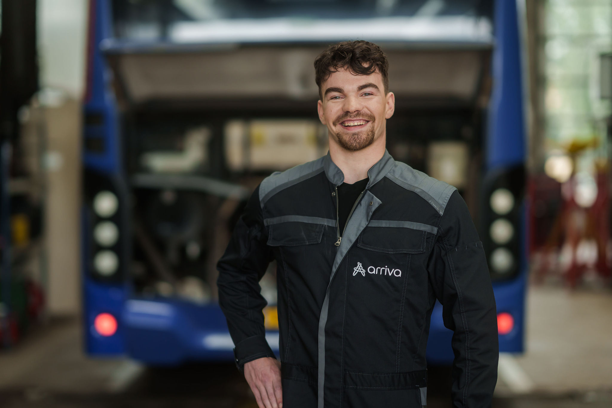 Een alchende monteur in Arriva-werkkleding staat in een garage, met een blauwe bus op de achtergrond.