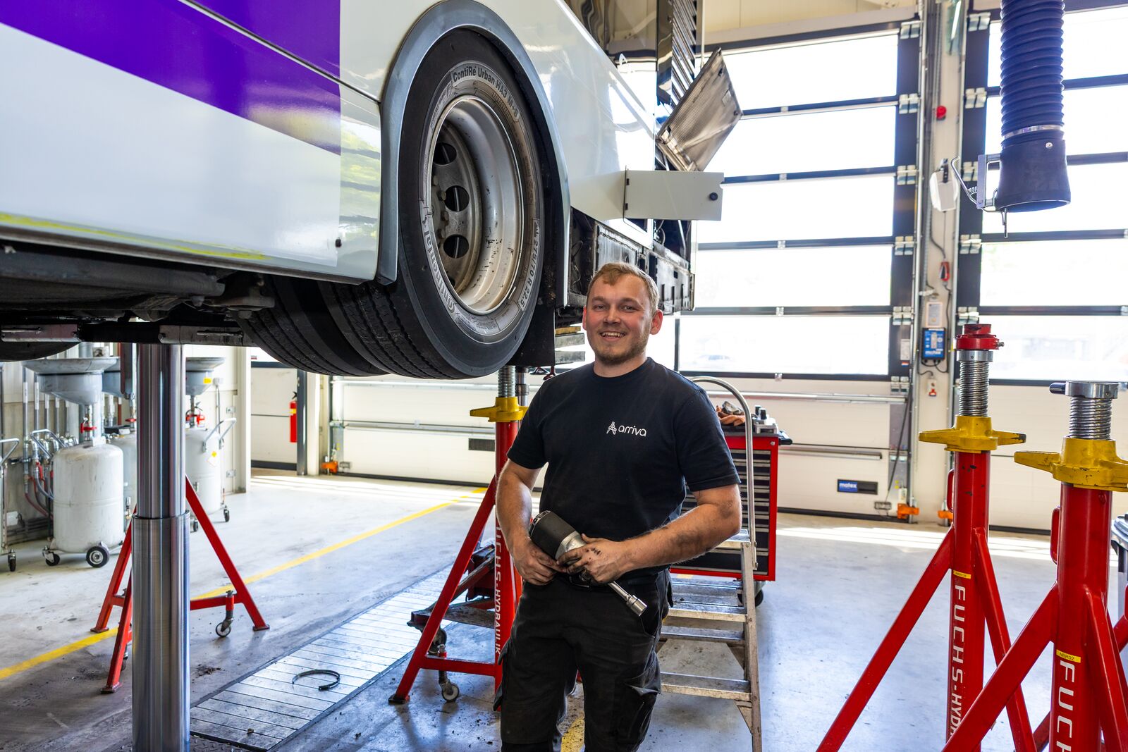 Monteur staat lachend bij wiel van een bus terwijl hij apparatuur vasthoudt.
