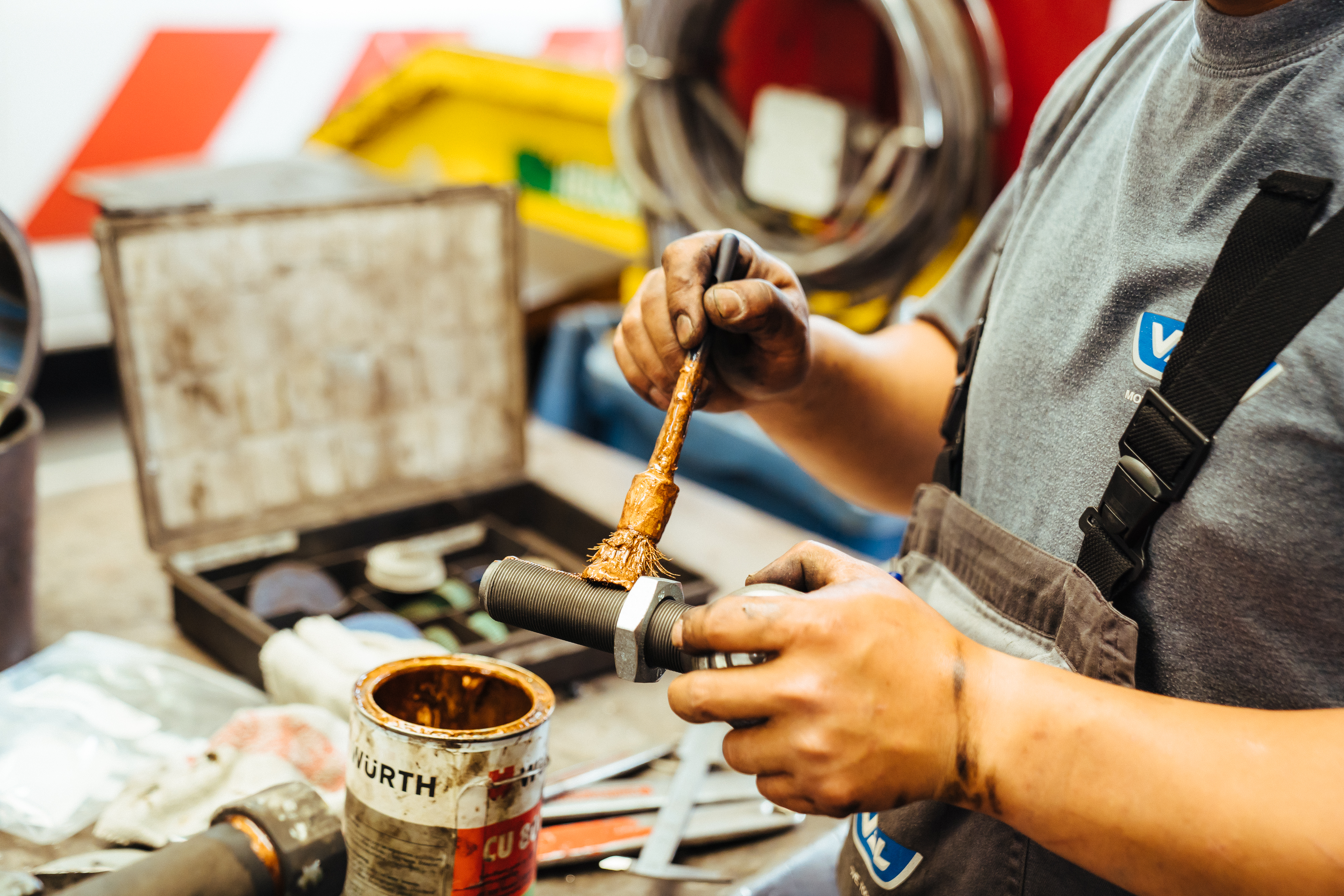 Monteur smeert een bout in met een kwast in een werkplaats met gereedschap op de achtergrond.