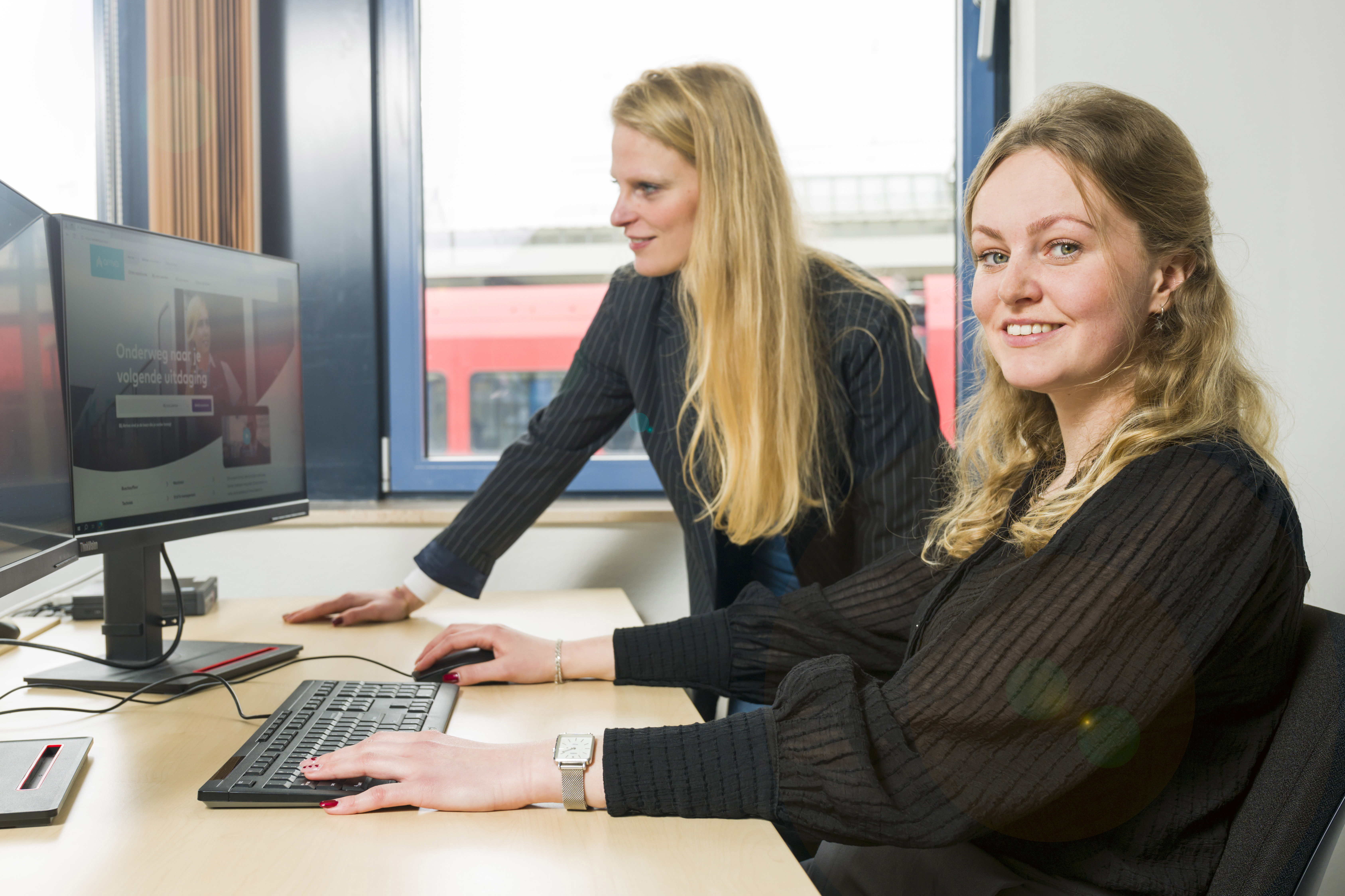 Twee vrouwelijke trainees, één zittend achter computer, ander staat ernaast, samen kijken ze naar computerscherm.  