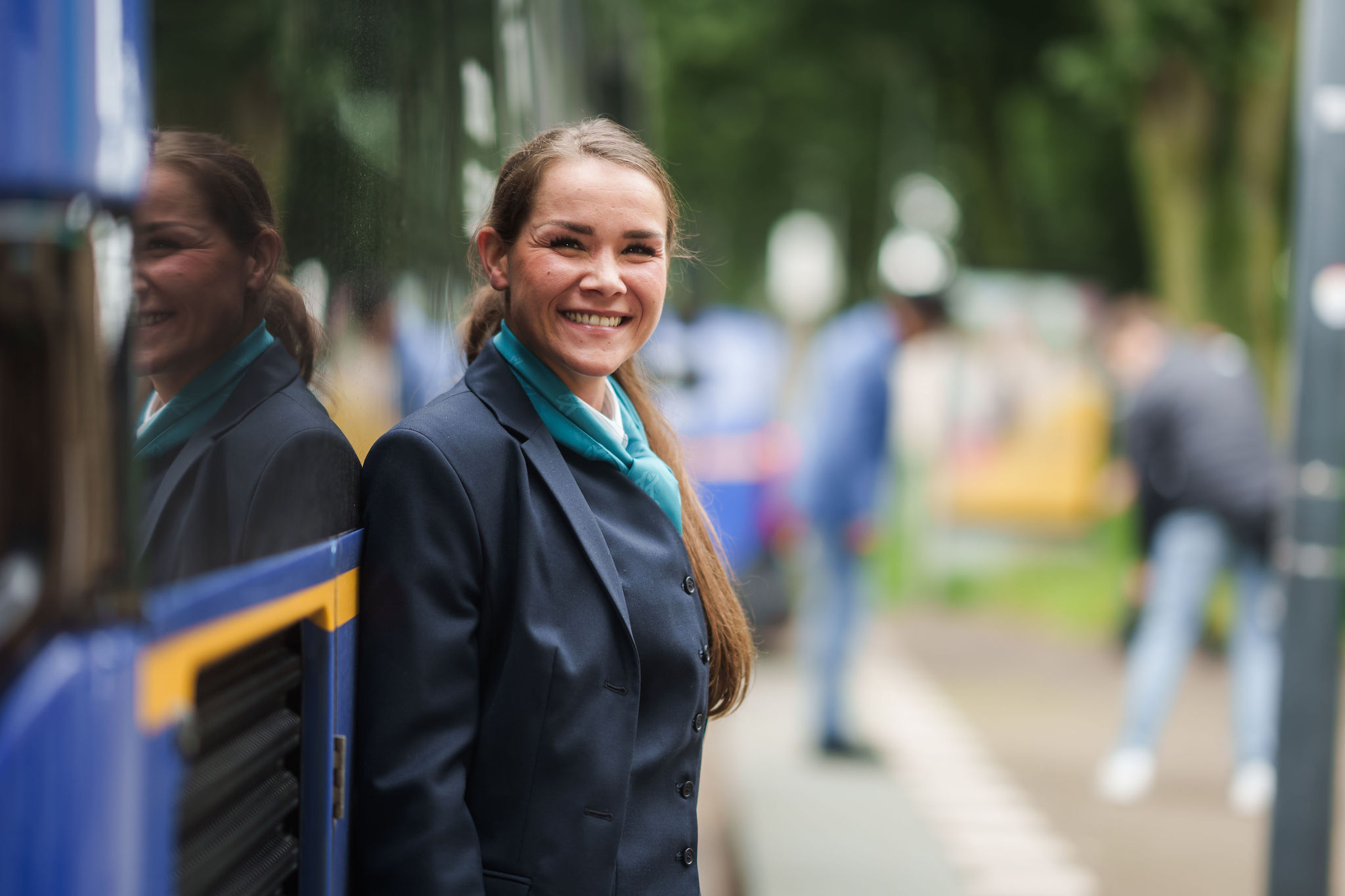 Buschauffeur van Arriva staat naast haar bus en ze glimlacht, je ziet haar weerspiegeling in de bus.