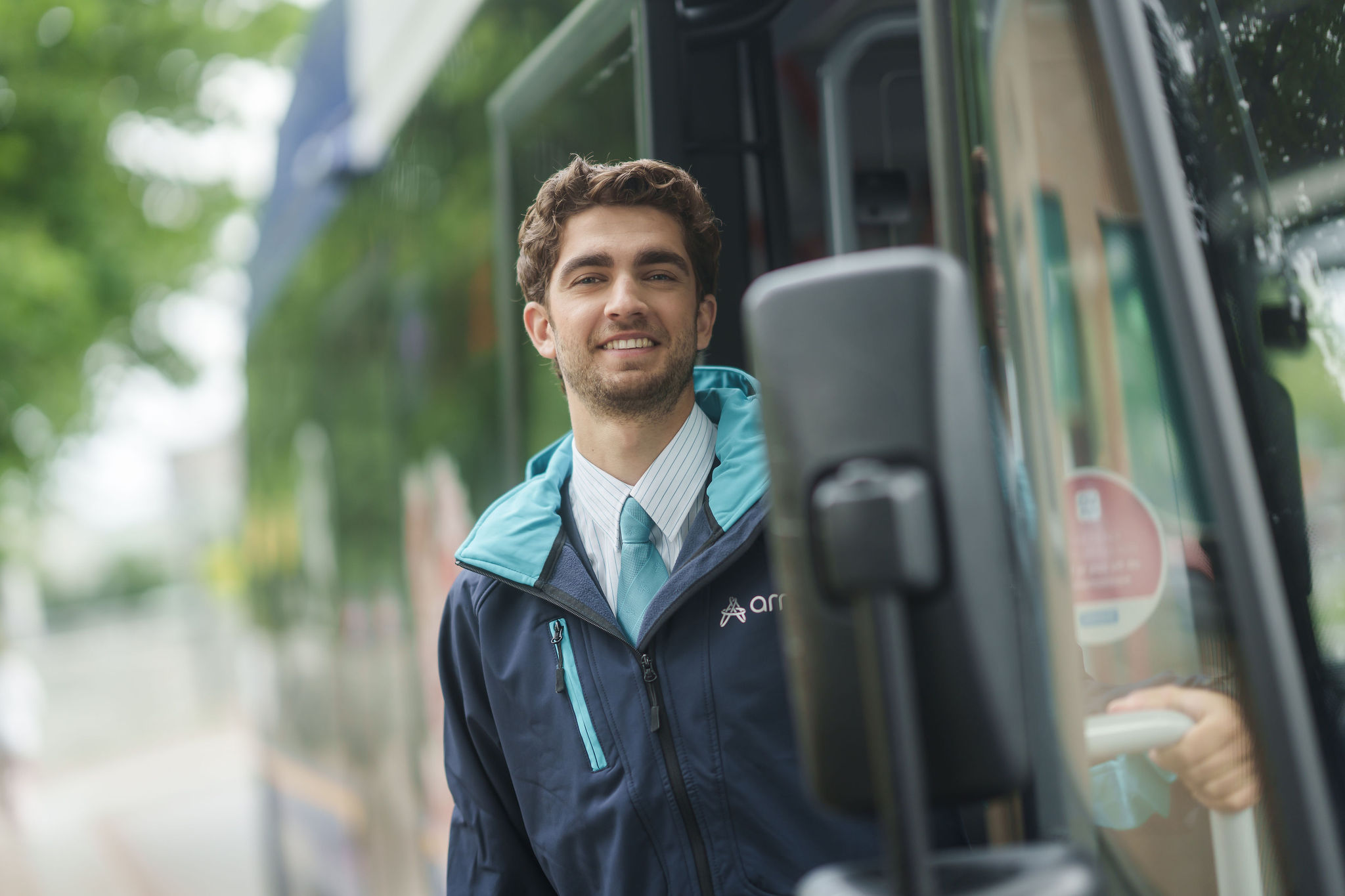 Een chauffeur van Arriva in uniform lacht naar de camera terwijl hij in de deuropening van zijn bus staat.