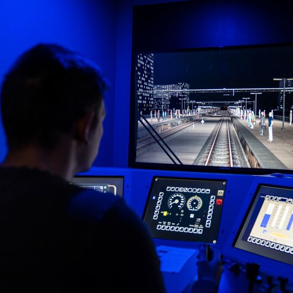 Een man zit in een treinsimulator, hij kijkt naar een groot scherm met daarop het spoor en heeft zijn handen op de stuurhendels liggen. Een man zit in een treinsimulator, hij kijkt naar een groot scherm met daarop het spoor en heeft zijn handen op de stuurhendels liggen.