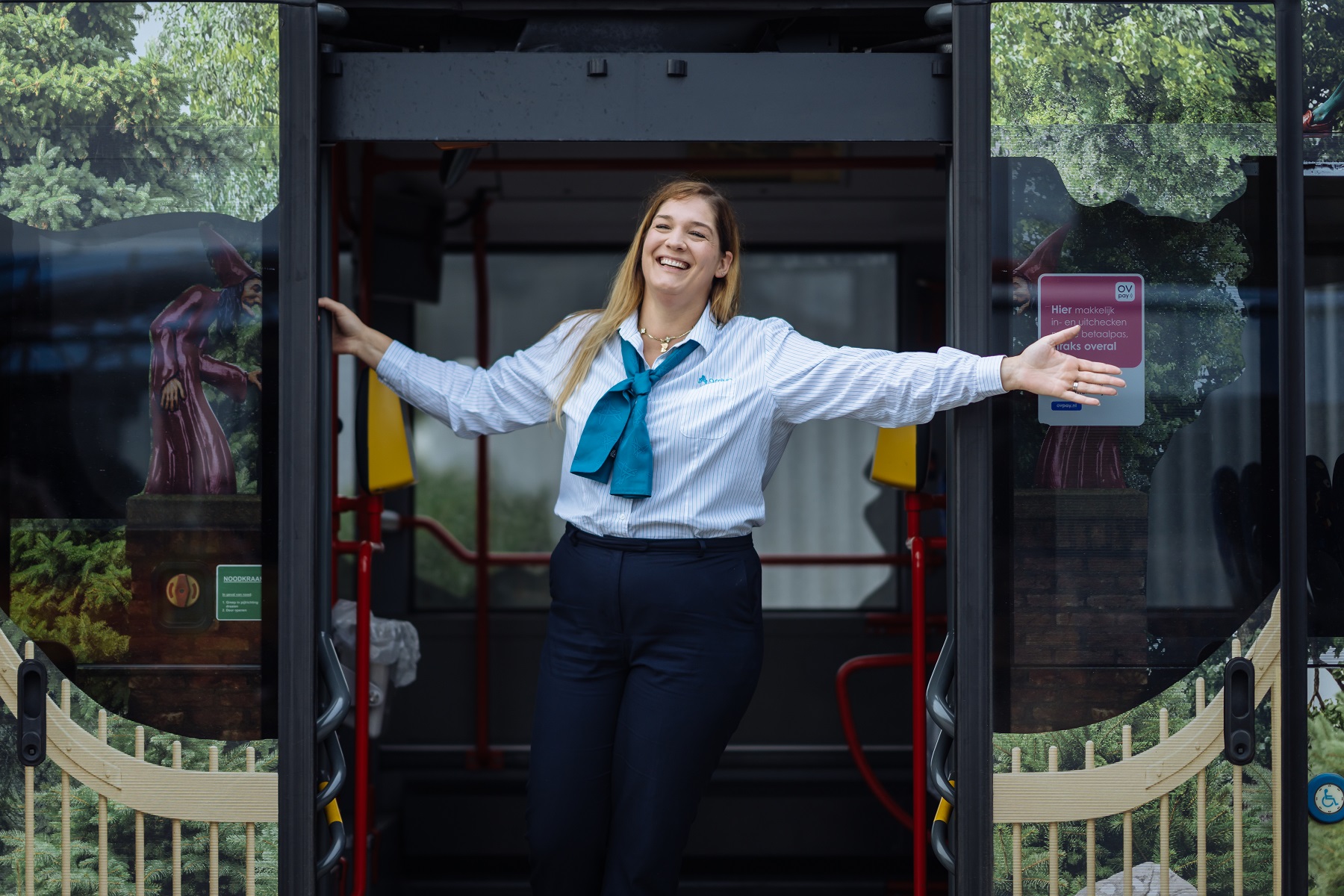 Buschauffeur van Arriva staat breed lachend met haar armen wijd in de deuropening van een bus.