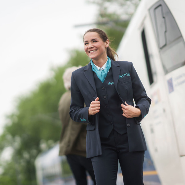 Een lachende machinist in Arriva-uniform staat buiten naast een trein, met bomen en een andere persoon op de achtergrond. Een lachende machinist in Arriva-uniform staat buiten naast een trein, met bomen en een andere persoon op de achtergrond.