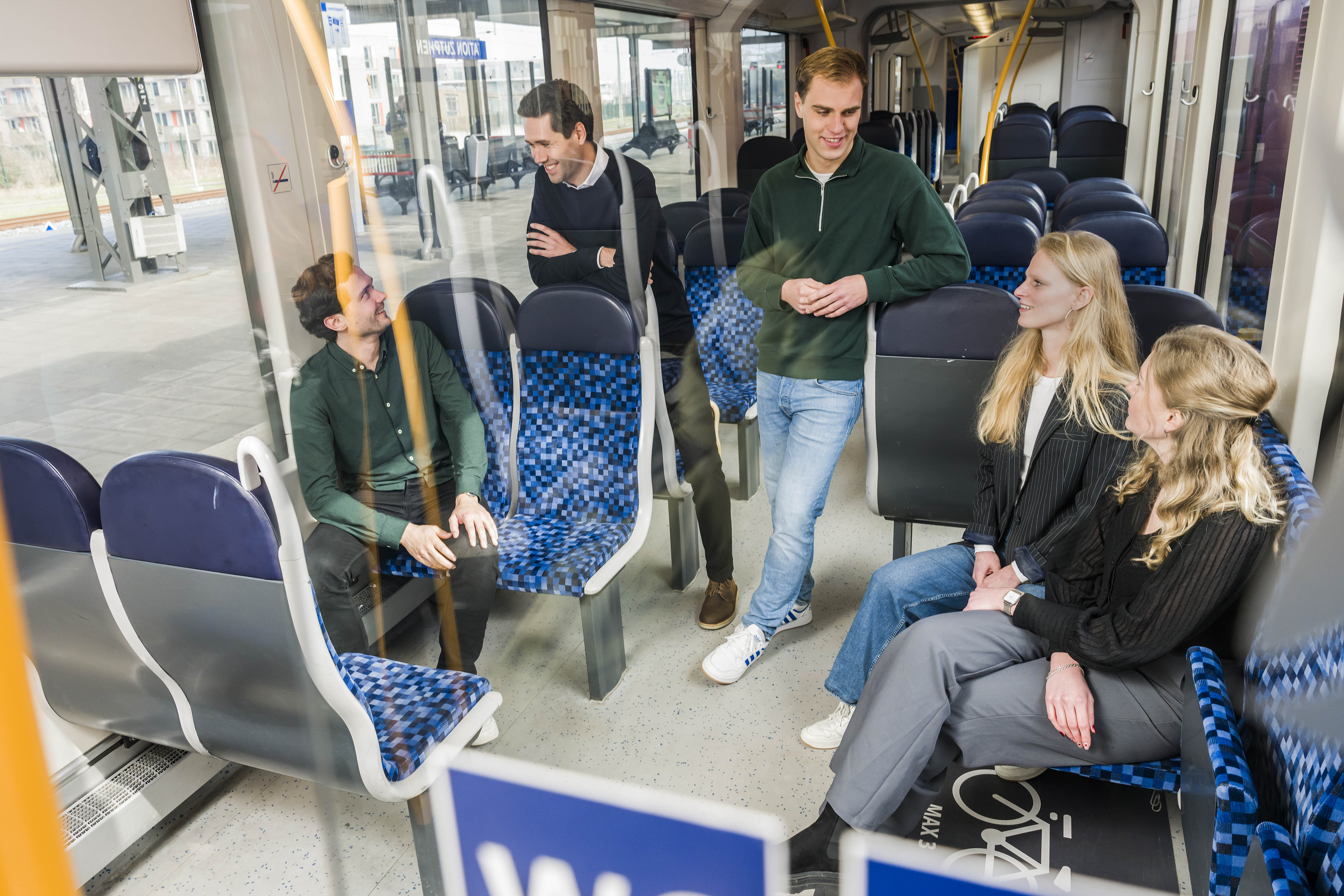 Vier trainees in gesprek in lege Arriva-trein, twee staan, twee zitten, ontspannen sfeer met natuurlijk licht door ramen.