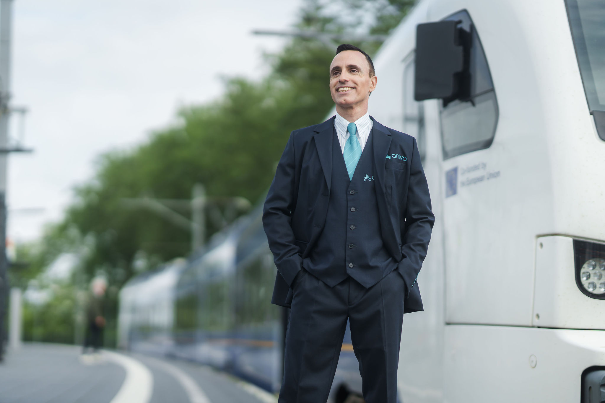 Machinist van Arriva glimlacht en kijkt in de verte met achter hem een trein.