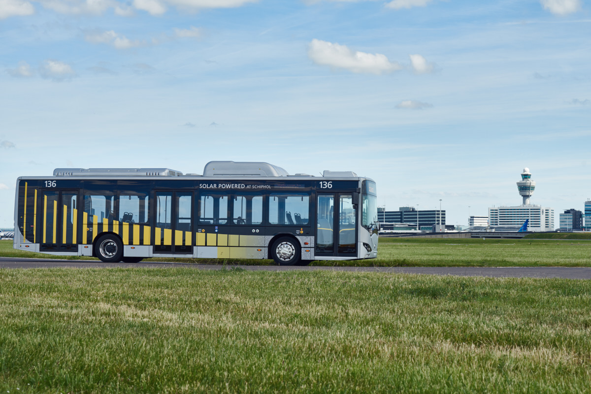 Elektrische Arriva-bus rijdt door een weiland onder een blauwe lucht met wolken. Op de achtergrond is Schiphol te zien.