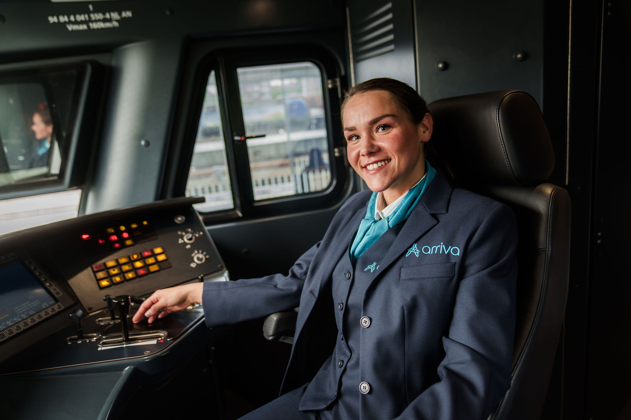 Een lachende machinist in Arriva-uniform zit in de bestuurderscabine van een trein en kijkt naar de camera.