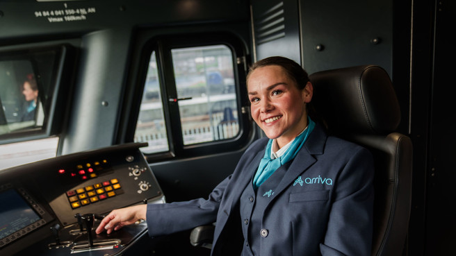 Een lachende machinist in Arriva-uniform zit in de bestuurderscabine van een trein en kijkt naar de camera. Een lachende machinist in Arriva-uniform zit in de bestuurderscabine van een trein en kijkt naar de camera.