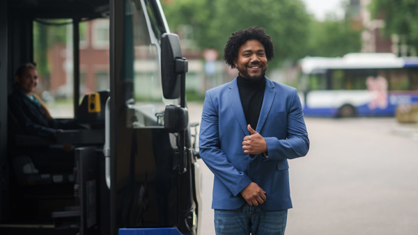 Een teammanager van Arriva in blauw pak staat lachend naast een bus. Een teammanager van Arriva in blauw pak staat lachend naast een bus.