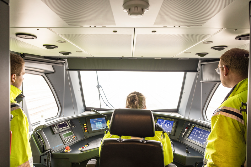 Drie Arriva-medewerkers, van achter gezien, in de cabine van een trein met zicht op spoor en bedieingspanelen.
