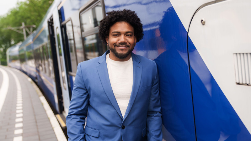 Een teammanager van Arriva staat in blauw pak en glimlachend bij een blauw-witte trein. Een teammanager van Arriva staat in blauw pak en glimlachend bij een blauw-witte trein.