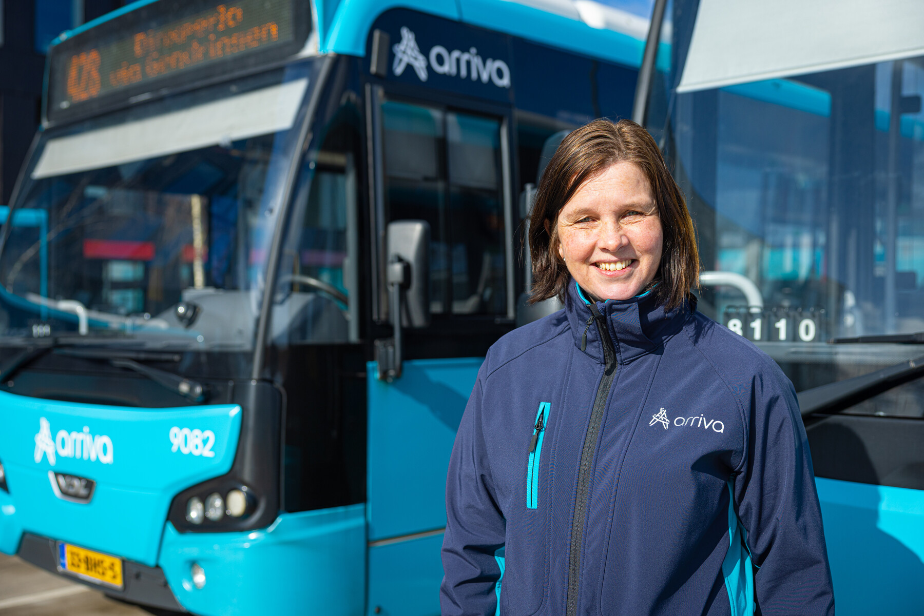 Trots glimlachende buschauffeur staand voor twee blauwe Arriva bussen van regio Oost.