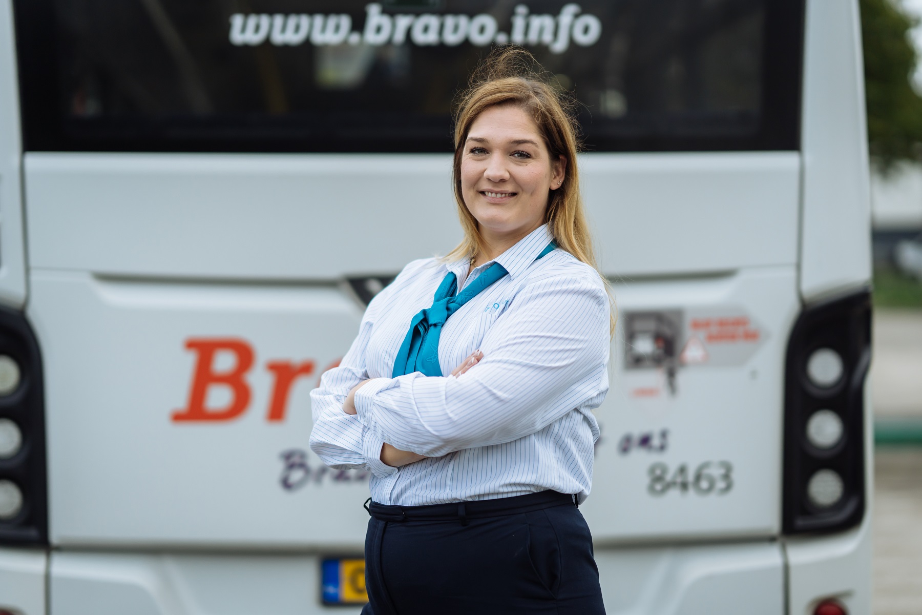 Buschauffeur van Arriva staat achter een een bus, lacht naar de camera en heeft haar armen over elkaar.