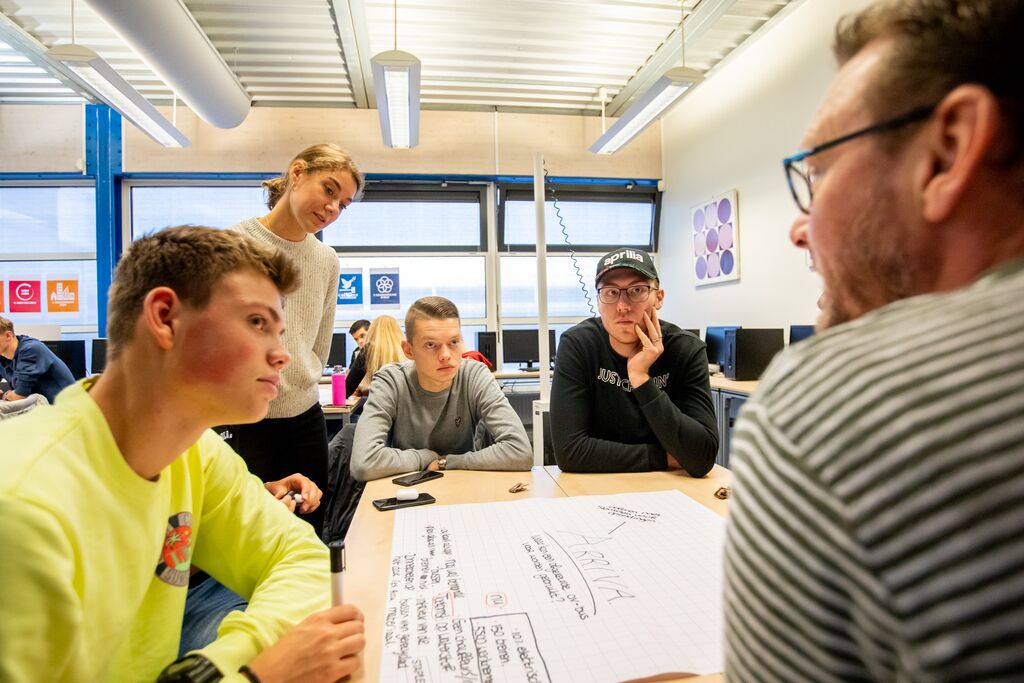 Een groep jongeren zit rond een tafel in een klaslokaal en kijken naar de leraar.