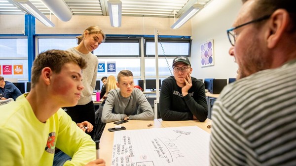 Een groep jongeren zit rond een tafel in een klaslokaal en kijken naar de leraar. Een groep jongeren zit rond een tafel in een klaslokaal en kijken naar de leraar.