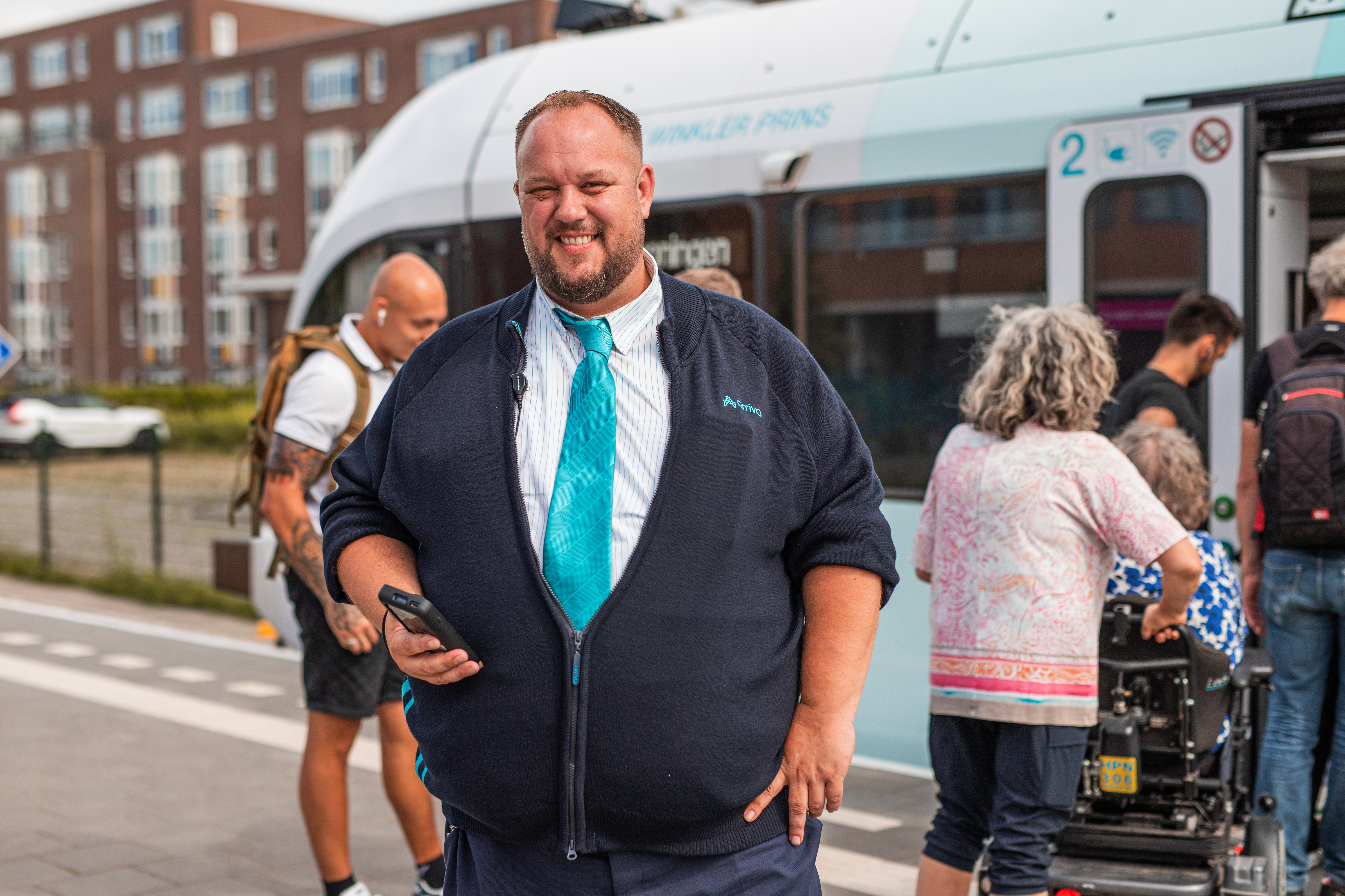 Steward Peter staat in zijn donkerblauwe Arriva vest en aquakleurige Arriva stropdas voor de trein. Achter hem stappen reizigers in. Hij gaat ze reisbewijzen controleren met het apparaat dat hij daarvoor in zijn werk gebruikt. 