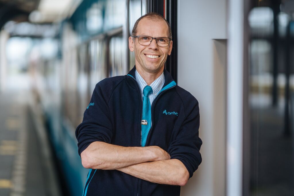 Een vrolijke man in een Arriva-uniform staat met zijn armen over elkaar en leunt tegen een trein, terwijl hij glimlacht.