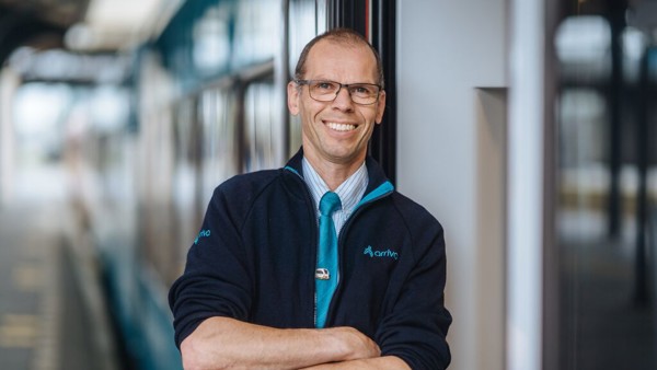 Een vrolijke man in een Arriva-uniform staat met zijn armen over elkaar en leunt tegen een trein, terwijl hij glimlacht. Een vrolijke man in een Arriva-uniform staat met zijn armen over elkaar en leunt tegen een trein, terwijl hij glimlacht.