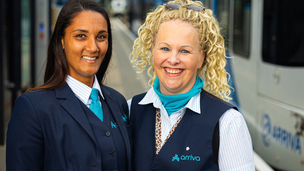 Twee vrouwelijke Stewards BOA-OV van Arriva in uniform staan glimlachend naast elkaar op een perron, met een trein op de achtergrond. Twee vrouwelijke Stewards BOA-OV van Arriva in uniform staan glimlachend naast elkaar op een perron, met een trein op de achtergrond.