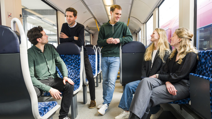 Vijf jonge trainees staan en zitten in treinwagon, levendig discussiërend, de binnenkant van de moderne Arriva-trein zichtbaar. Vijf jonge trainees staan en zitten in treinwagon, levendig discussiërend, de binnenkant van de moderne Arriva-trein zichtbaar.