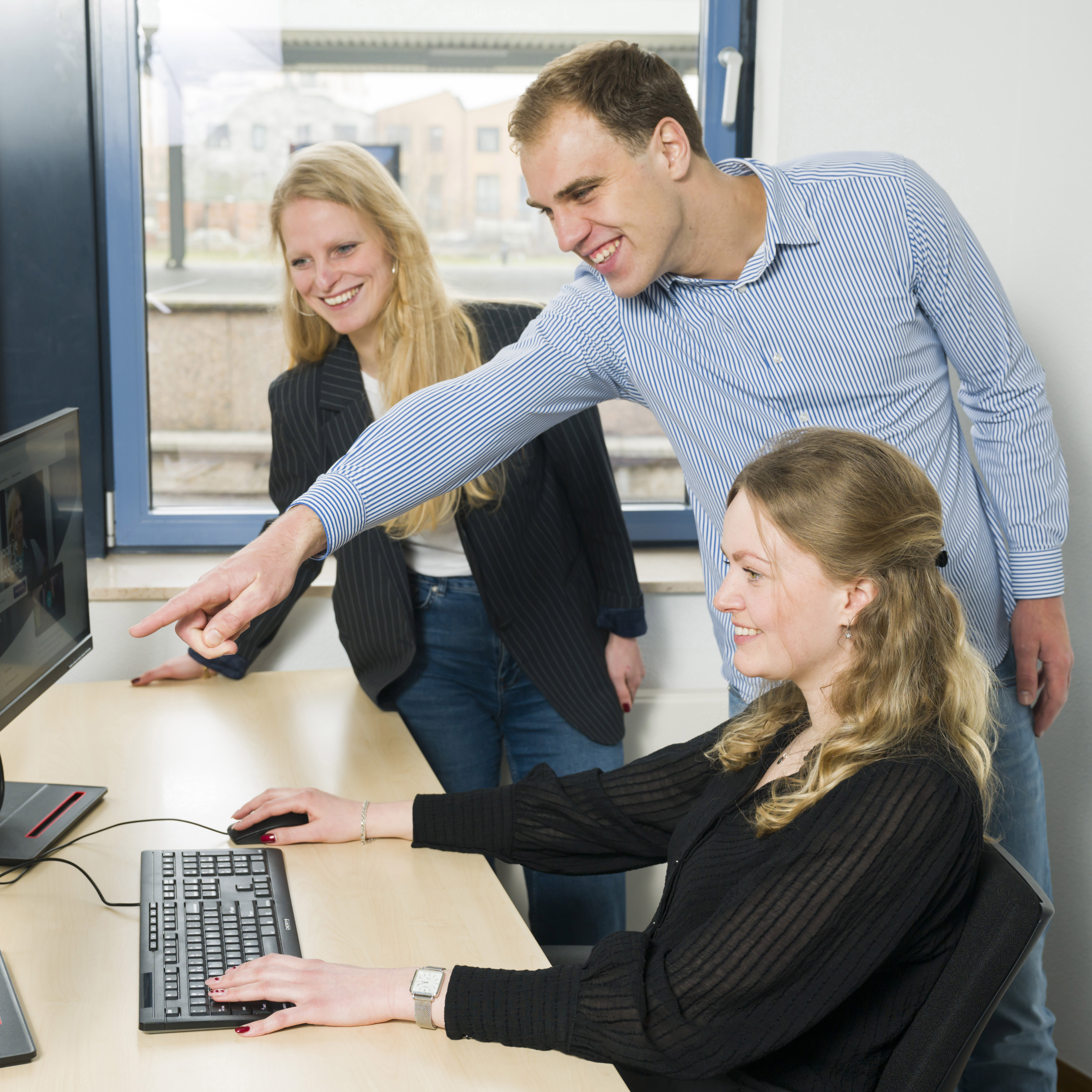 Twee vrouwen en een man werken lachend samen op kantoor, man staat en wijst naar computerscherm, vrouwen zitten en kijken aandachtig.