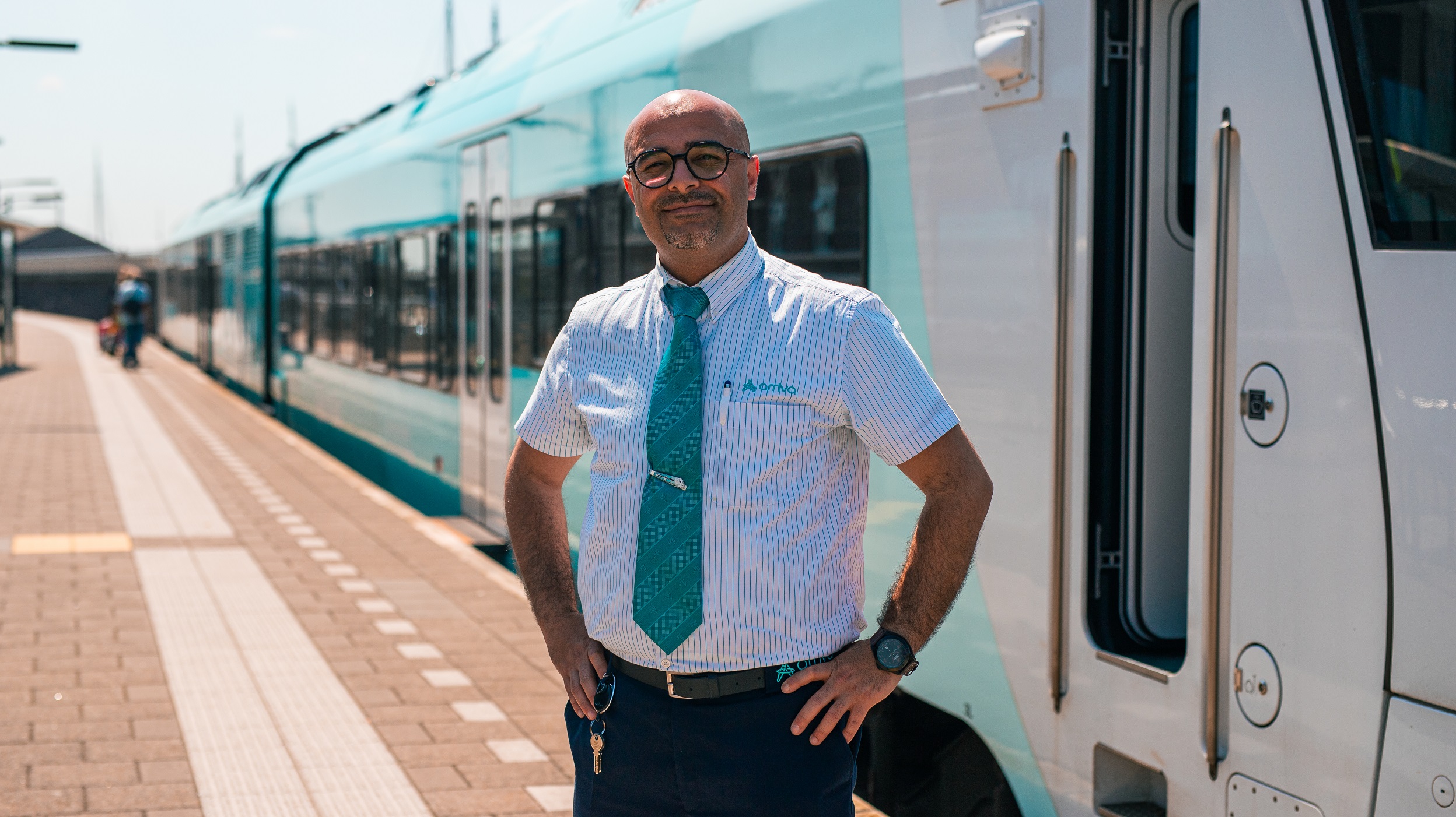 Een lachende machinist in Arriva-uniform staat voor de trein en kijkt in de camera. 