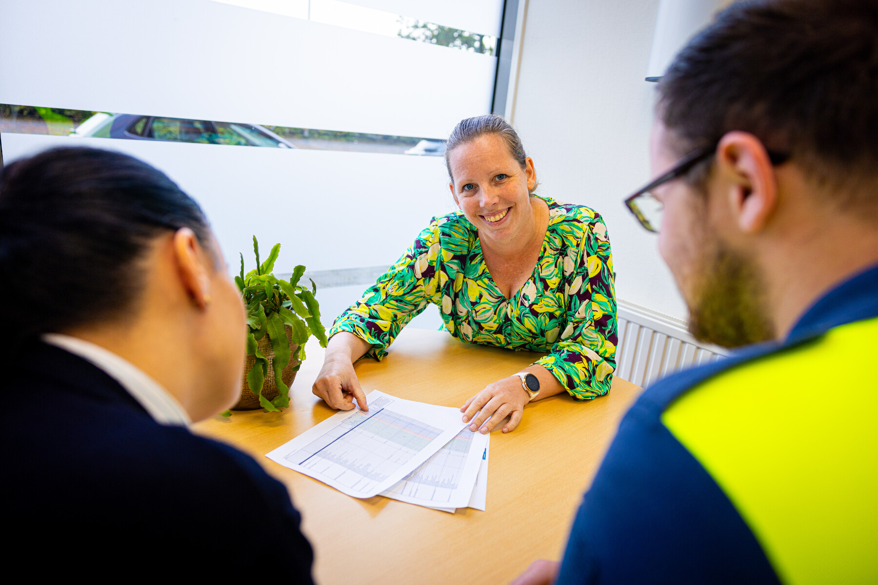 Teammanager Irene zit lachend aan tafel en reikt twee collega's papieren aan.