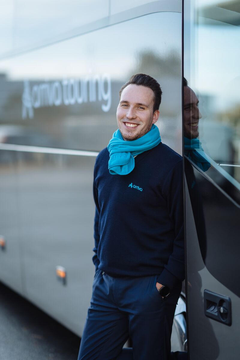 Touringcarchauffeur Benjaming staat lachend naast zijn touringcar.