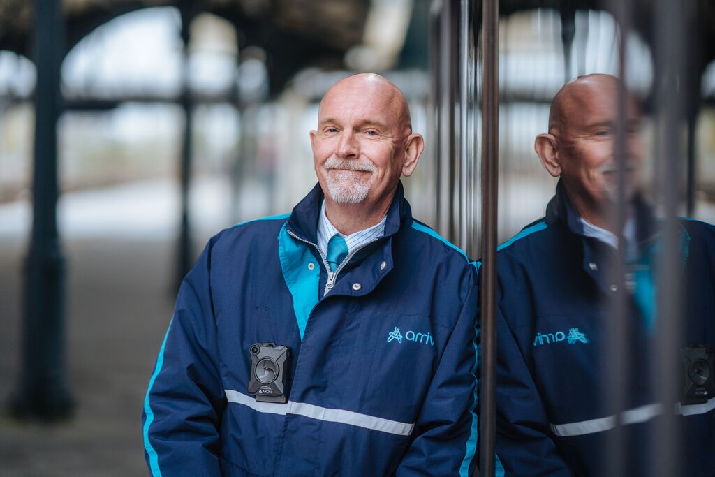 Een mannelijke conducteur met een Arriva-jas leunt ontspannen tegen de zijkant van een Arriva-trein.