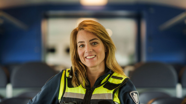 Medewerker service & veiligheid van Arriva staat in haar uniform in een trein tussen de stoelen en lacht. Medewerker service & veiligheid van Arriva staat in haar uniform in een trein tussen de stoelen en lacht.